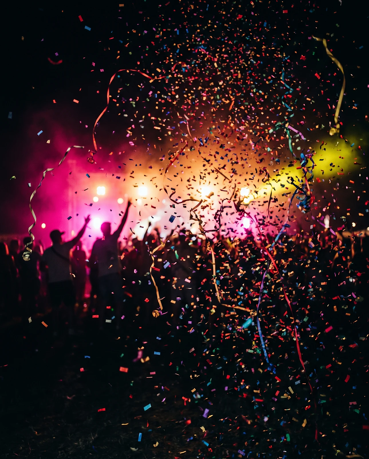 Wolk van gekleurde confetti en linten boven een donkere dansvloer, met op de achtergrond silhouetten van feestgangers met de armen omhoog in roze en geel licht